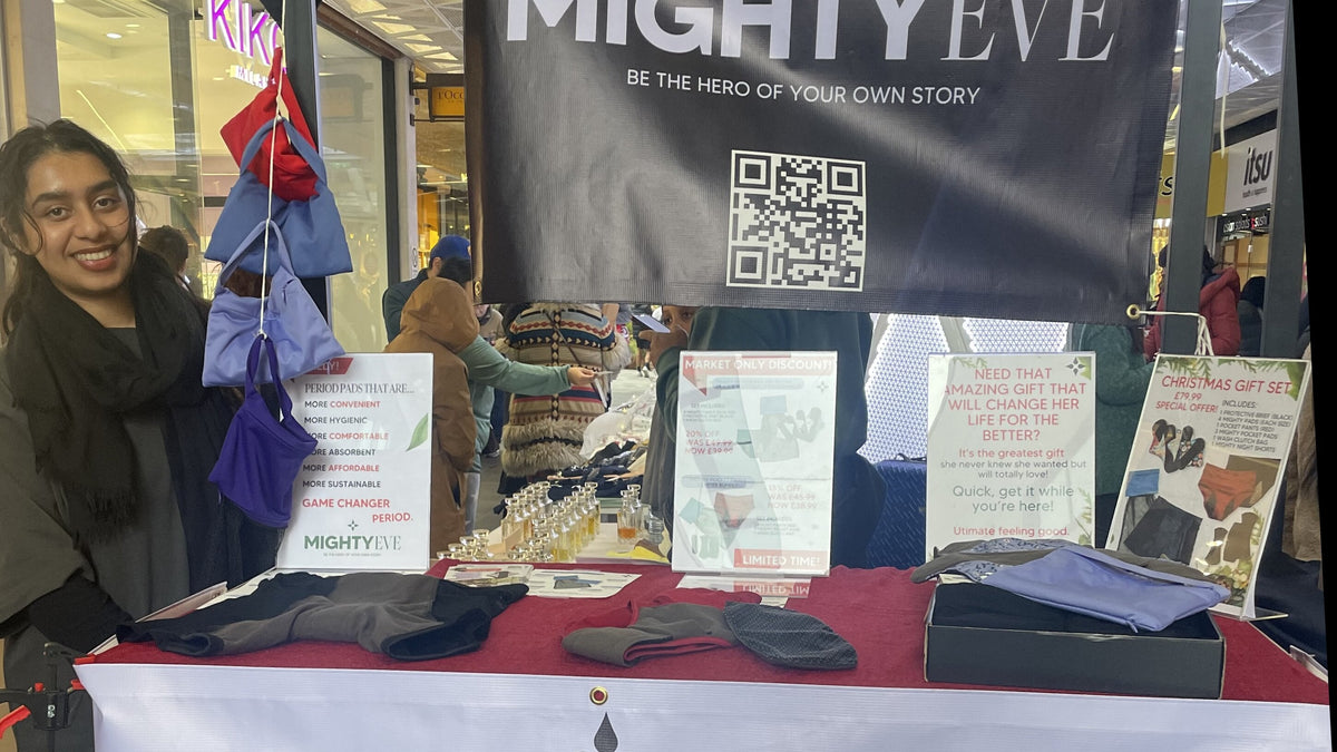 A smiling woman behind the Mighty Eve stall at Angel Central, with folded products and a gift box on the table, drawstring pouches above, a Christmas Gift Set sign, and the MightyEve banner with QR code behind her.