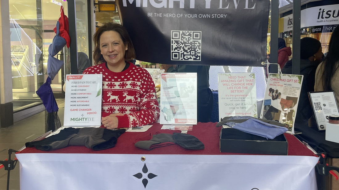 A smiling woman in a red Christmas jumper behind the Mighty Eve stall at Angel Central, with period pants and shorts on the table, a Christmas Gift Set special offer sign, the MightyEve logo banner, and a QR code behind her.