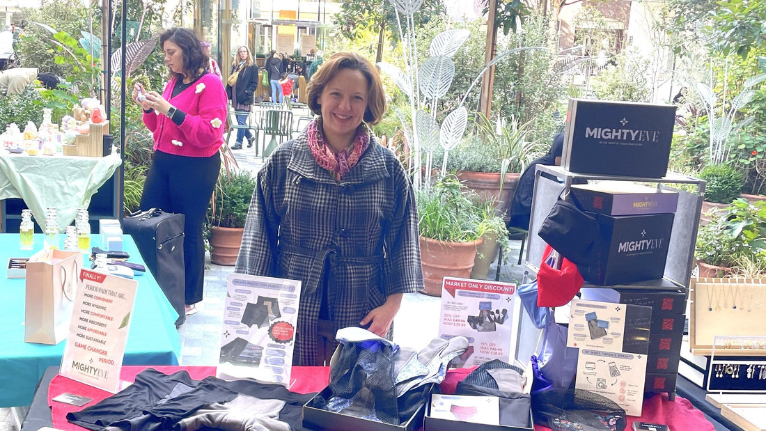Mighty Eve founder Sara smiling behind the MightyEve stall at Angel Central sustainability market, with period pants, product boxes, and promotional signage displayed on the table.