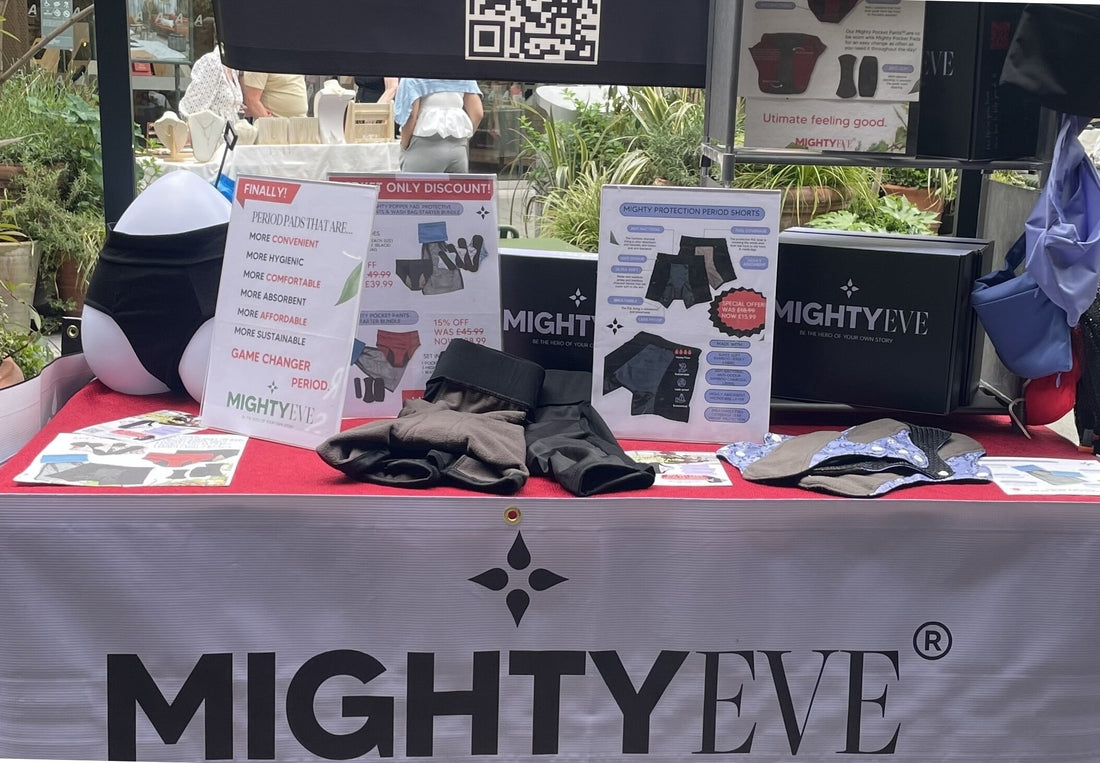 MightyEve market stall at Angel Central: a black overhead banner with a QR code and product graphics, branded boxes reading 'Be the hero of your own story', period pads and shorts signage, and a grey tablecloth with the MightyEve wordmark.