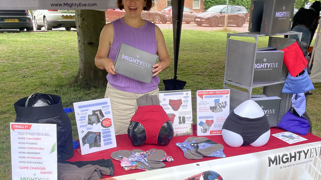 A smiling woman holding a grey MightyEve gift box at the Mighty Eve stall, with period pants, pads and shorts displayed on a red-clothed table and a Market Only Discount sign.