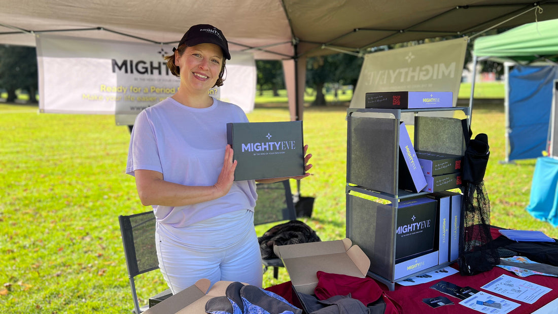 Sara from Mighty Eve smiling under a gazebo at Harpenden Sustainability Market, holding a branded box, with period pants stock on the stall table beside her.