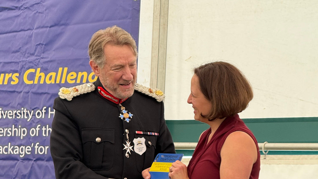 The Lord-Lieutenant of Hertfordshire, in ceremonial dress uniform, shaking hands with a Mighty Eve representative and presenting the Entrepreneurs Challenge winner's plaque, with the event's banner visible behind them.
