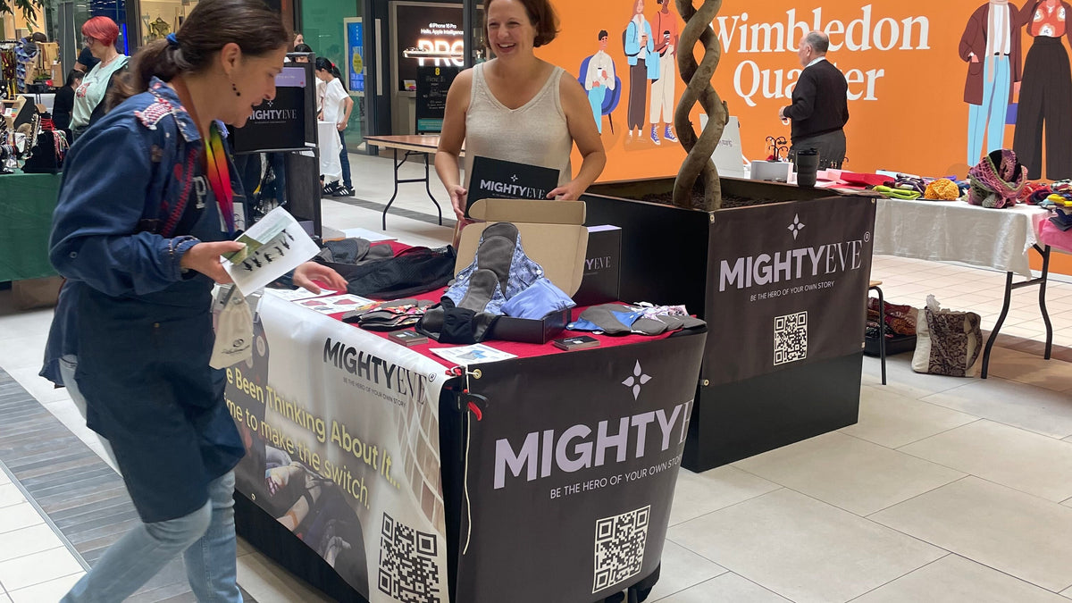 The Mighty Eve market stall inside Wimbledon Quarter shopping centre, with period pants and pouches on the table, a customer browsing, MightyEve branding, QR codes, and the Wimbledon Quarter sign behind.