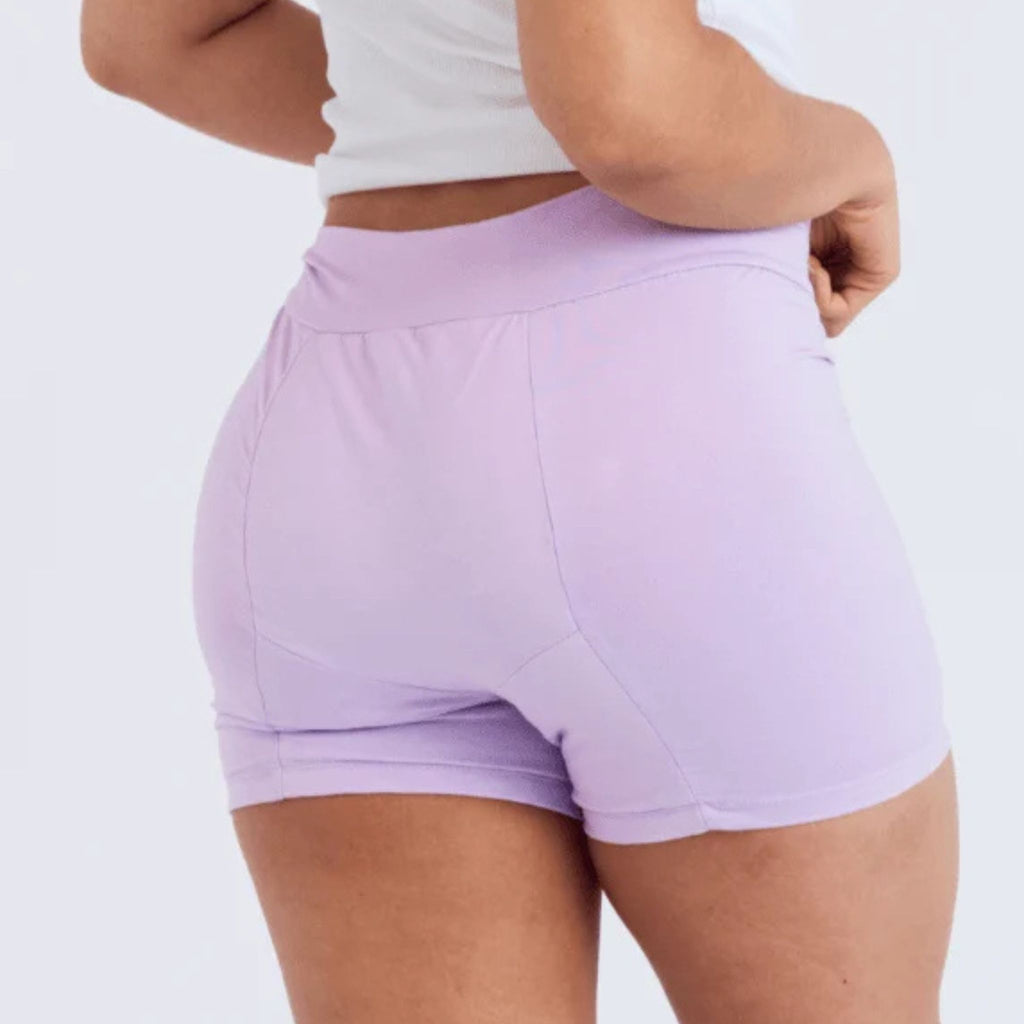 A close-up back view of a person wearing high-waisted light purple period shorts and a white cropped tank top against a plain light background.